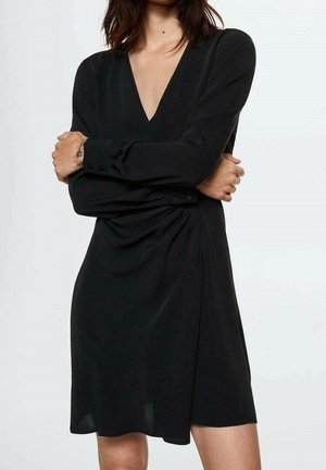 Donna con le braccia incrociate che indossa un vestito nero a maniche lunghe con scollo a V profondo e polsini abbottonati, in piedi contro uno sfondo chiaro e semplice.