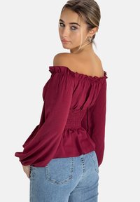 Burgunderfarbene Off-Shoulder-Bluse mit geraffter Taille, gepufften Ärmeln und strukturiertem Stoff, kombiniert mit hellen, eng anliegenden Jeans.