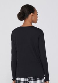 Černá dlouhá halenka s lemováním ve vlnkách, hladká textilie a volný střih, doplněná kostkovanými pyžamovými kalhotami.