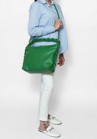 Sac à main en cuir vert avec une anse tressée, de forme rectangulaire et des accents en métal doré. Présente une texture lisse et une bandoulière ajustable.