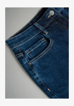 Tamsiai mėlynos džinsinės kelnės su priekiniais kišeniais, kontrastinga auksine siūle ir metaliniu mygtuku. Lygi tekstūra su lengvu blizgesiu.