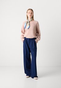 Roze donzige trui, marineblauwe wijde broek en een patroon sjaal om de nek. Crèmekleurige schoenen, minimale accessoires, effen achtergrond.
