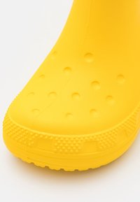 Parte frontal de una bota de goma amarilla brillante para la lluvia con patrones circulares elevados y borde de suela texturizado sobre un fondo blanco.