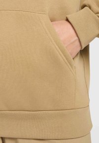 Brun bomullsgenser med lomme, ribbestrikkede mansjetter og sømløs kant. Har en myk tekstur og en avslappet passform.