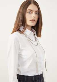 Witte button-up blouse van textuurstof, gecombineerd met een zwarte geruite rok. Een zilveren ketting met franjes accentueert de outfit.