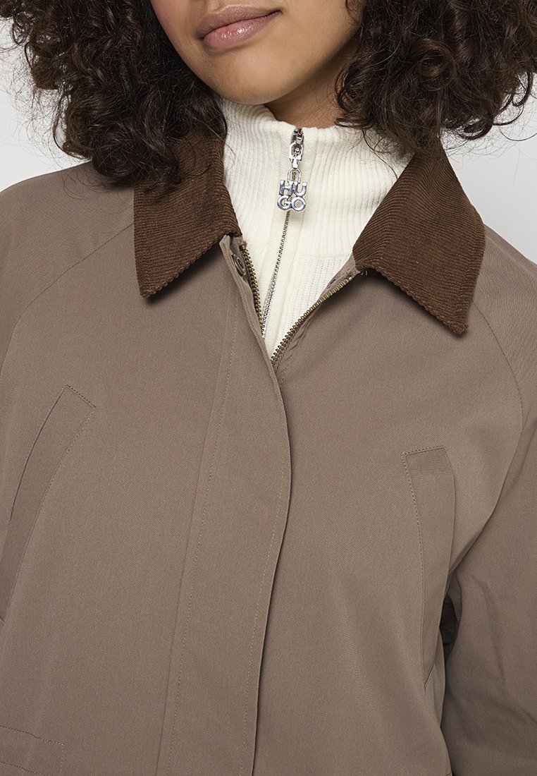 Bruine buitenjasje met een corduroy kraag. Lijkt gestructureerd met zijzakken en een ritssluiting, gedragen boven een wit geribbeld innerlijk topje.