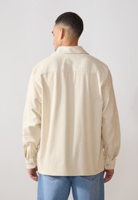 Chemise en velours côtelé beige avec un col pointu, des manches longues et des poignets boutonnés. Des lignes verticales texturées traversent tout le tissu.