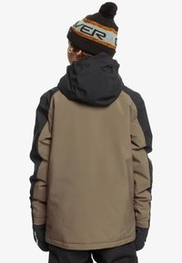Braune und schwarze Winterjacke mit Kapuze, mit glatter Textur und elastischen Bündchen, getragen mit einer schwarzen Strickmütze mit orangefarbenen Streifen.
