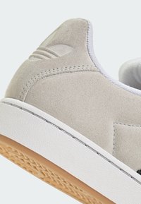 Svetlosivá semišová teniska s bielou gumovou podrážkou a textúrovanými detailmi. Obsahuje logo na päte a polstrovaný golier pre pohodlie.