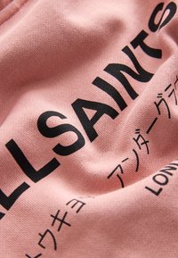 Makro záber na ružovú látku s čiernym textom "ALLSAINTS" a vytlačenými japonskými znakmi.