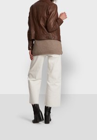 Hnědá kožená bunda přes béžový svetr, spárovaná s bílými širokými kalhotami a černými vysokými kotníčkovými botami. Viditelné švy na bundě.