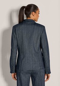 Vrouw met donker haar in een paardenstaart, gekleed in een strakke, donkere streepjumpsuit, met haar rug naar de camera gekeerd tegen een effen lichte achtergrond.