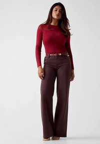 Haut rouge à manches longues côtelé avec un décolleté transparent, associé à un pantalon large marron taille haute et une ceinture assortie.