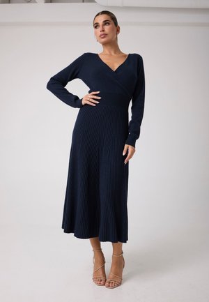 Femme portant une longue robe en maille côtelée bleu foncé à manches longues et des sandales à talons hauts beiges, debout devant un fond blanc uni.