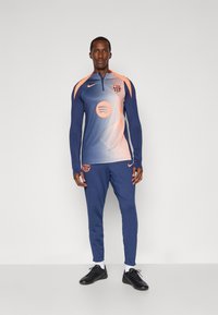 Ensemble de vêtements de sport Nike en bleu marine et orange, comprenant un haut zippé avec un motif dégradé, et un pantalon ajusté avec le logo du FC Barcelone.