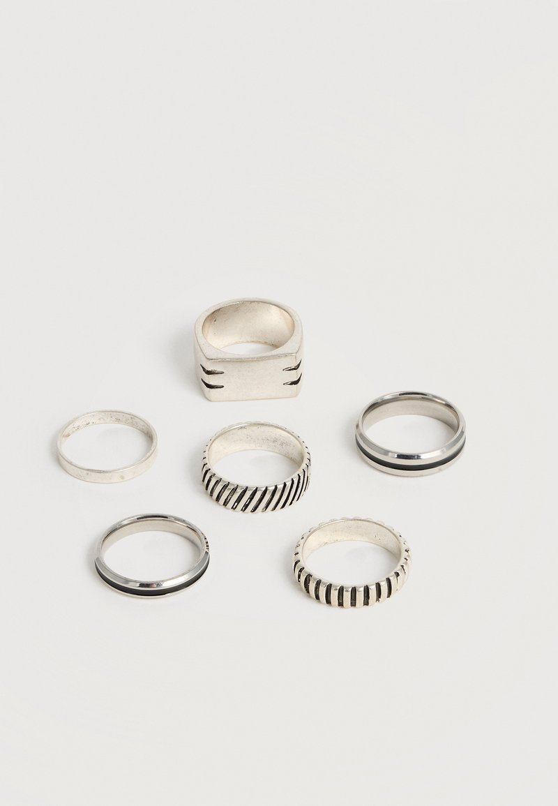 Έξι ασημένια δαχτυλίδια με διαφορετικά σχέδια, περιλαμβάνονται λείες ζώνες, ραβδώσεις υφής και ένα παχύ τετράγωνο δαχτυλίδι, τοποθετημένα πάνω σε μια απλή λευκή επιφάνεια.