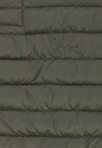 Dunkelgrüner, gestepptes Gewebe mit horizontalen Nähten und gesticktem Text "SUB LEVEL" in passendem grünen Faden.