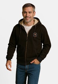 Zwarte zip-up hoodie met een tan fleece-voering aan de binnenkant, voorzien van decoratieve trekkoorden en voorzakken. Ggedragen over een wit shirt.