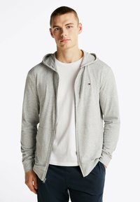 Sudadera con cremallera en color gris, fabricada con un material ligero y capucha con cordón. Camiseta blanca debajo. Presenta un pequeño logo en el pecho.