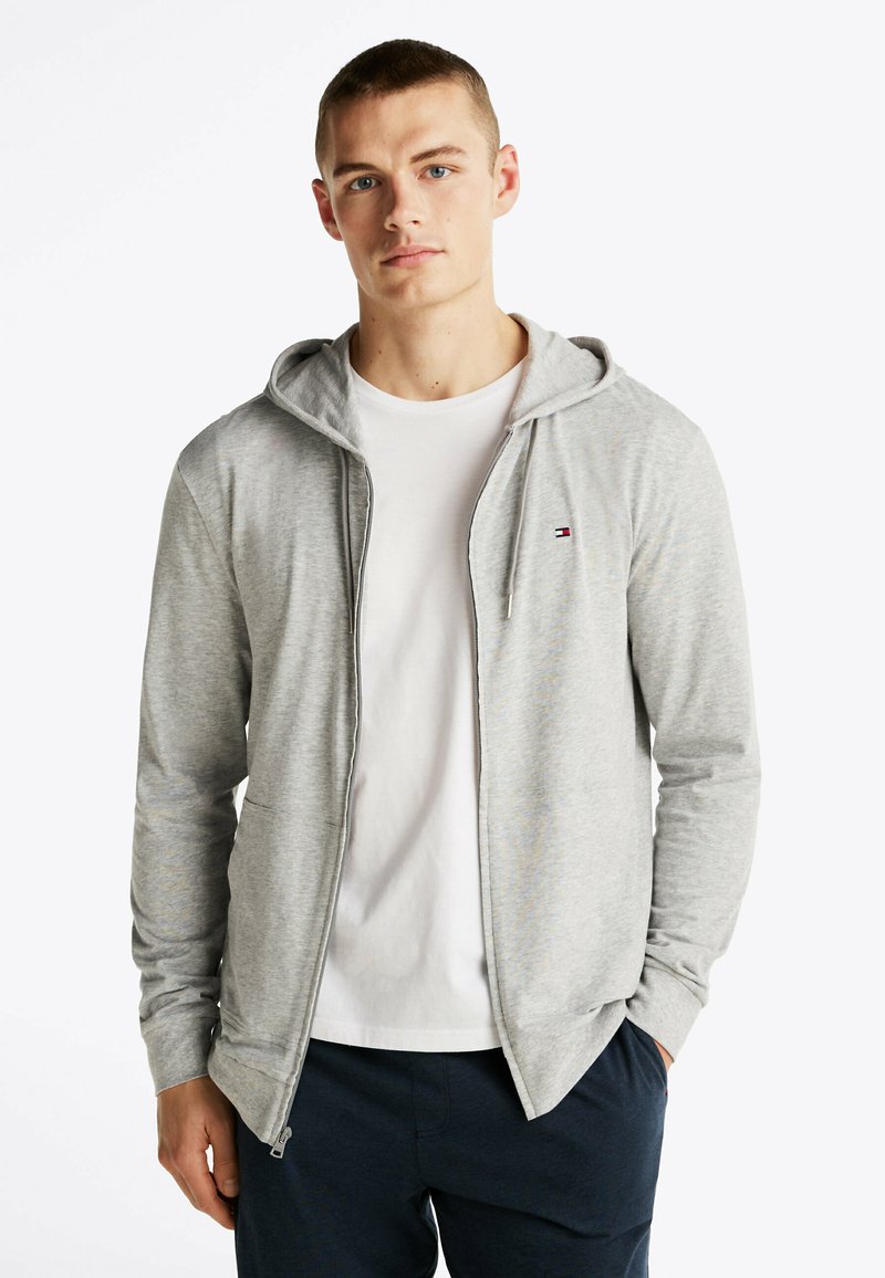 Sudadera con cremallera en color gris, fabricada con un material ligero y capucha con cordón. Camiseta blanca debajo. Presenta un pequeño logo en el pecho.