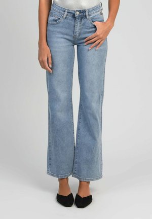 Femme portant un jean évasé taille haute bleu clair avec des poches avant et des chaussures noires à bouts pointus, se tenant devant un fond uni.