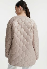 Gequilte buitenjack in lichtbeige, met een ronde halslijn, lange mouwen en een zachte, gladde textuur met verhoogde ovale patronen.