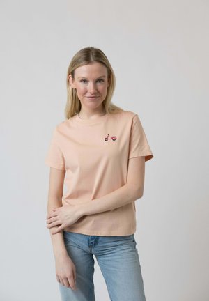 Junge Frau in einem pfirsichfarbenen T-Shirt mit kleinem pinkfarbenen Roller-Aufdruck auf der Brust, trägt hellblaue Jeans und steht vor einem einfarbigen hellen Hintergrund.
