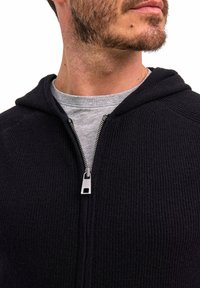 Schwarzer, gerippter Strick-Hoodie mit Reißverschluss und silbernen Beschlägen, über einem grauen Shirt getragen. Keine Muster oder auffälligen Akzente sichtbar.