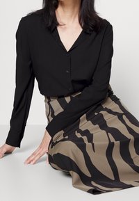 Schwarze Hemdbluse mit langen Ärmeln, kombiniert mit einem beigen Rock, der ein schwarzes Tigerstreifenmuster aufweist. Weicher Stoff mit glatter Textur.