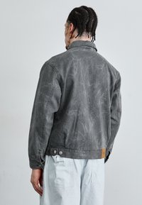 Gray denim jacket with a textured finish, featuring a collar, long sleeves, and snap button closure. Includes a brown leather accent at the hem.