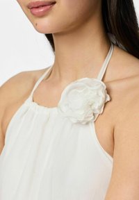 Haut halter blanc en tissu léger et fluide. Présente un décolleté froncé et un accent floral texturé blanc sur l'épaule.