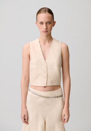 Mujer de piel clara y cabello recogido en un moño bajo, vestida con un chaleco sin mangas beige abotonado y una falda a juego con un cinturón decorativo de cadena plateada.