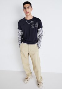 Chemise à rayures bleue marine à manches longues avec le numéro "24" imprimé ; pantalons beiges ; chaussures de couleur claire avec des découpes.