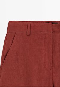 Pantalon en lin couleur rouille avec devant plat, passants pour ceinture, poches latérales, texture lisse et coupe ajustée.