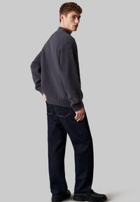 Maglione grigio scuro a maglia con colletto alto, polsini e orlo a costine, abbinato a jeans blu scuro e scarpe nere, visto da dietro.