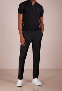Schwarzes Poloshirt mit kontrastierendem Kragen, kombiniert mit maßgeschneiderten schwarzen Hosen und weißen Sneakern. Verfügt �über ein goldenes Logo und seitliche Akzente.