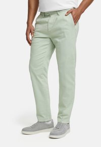 Light green, tapered chinos with a smooth texture, belt loops, side pockets, and contrasting gray sneakers, set against a plain background.
