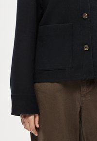 Casaco azul-marinho com colarinho redondo, apresentando um bolso no peito e botões de latão. Tecido com textura suave, combinado com calças castanhas.