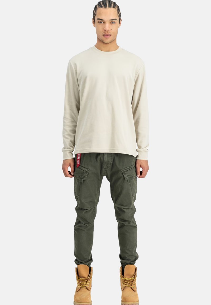 Camicia beige a maniche lunghe, pantaloni cargo verdi con tasche e stivali color sabbia. Vestibilità casual con design rilassato. I pantaloni presentano un'etichetta laterale con il marchio.