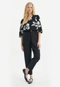 Zwarte bloes met bloemenmotief en witte patronen, opgeblazen mouwen en een V-hals. Zwarte tapse broek met knopen in de taille. Zwarte strap schoenen.