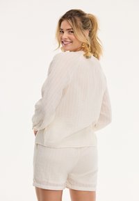 Blusa de lino blanca con mangas largas, patrón texturizado y corte holgado, combinada con pantalones cortos blancos. Destacado acento de botón dorado en los pantalones cortos.