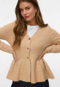 Trikotažna cardigan u svijetloj bež boji s V-izrezom, detaljem gumba i proširenim peplum rubom. Mekana tekstura s rebrastim rukavima i tijelom.