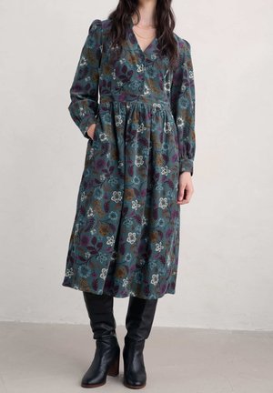 Femme portant une robe fleurie à manches longues et col en V avec des poches et des bottes noires à talons, debout à l’intérieur contre un mur uni.