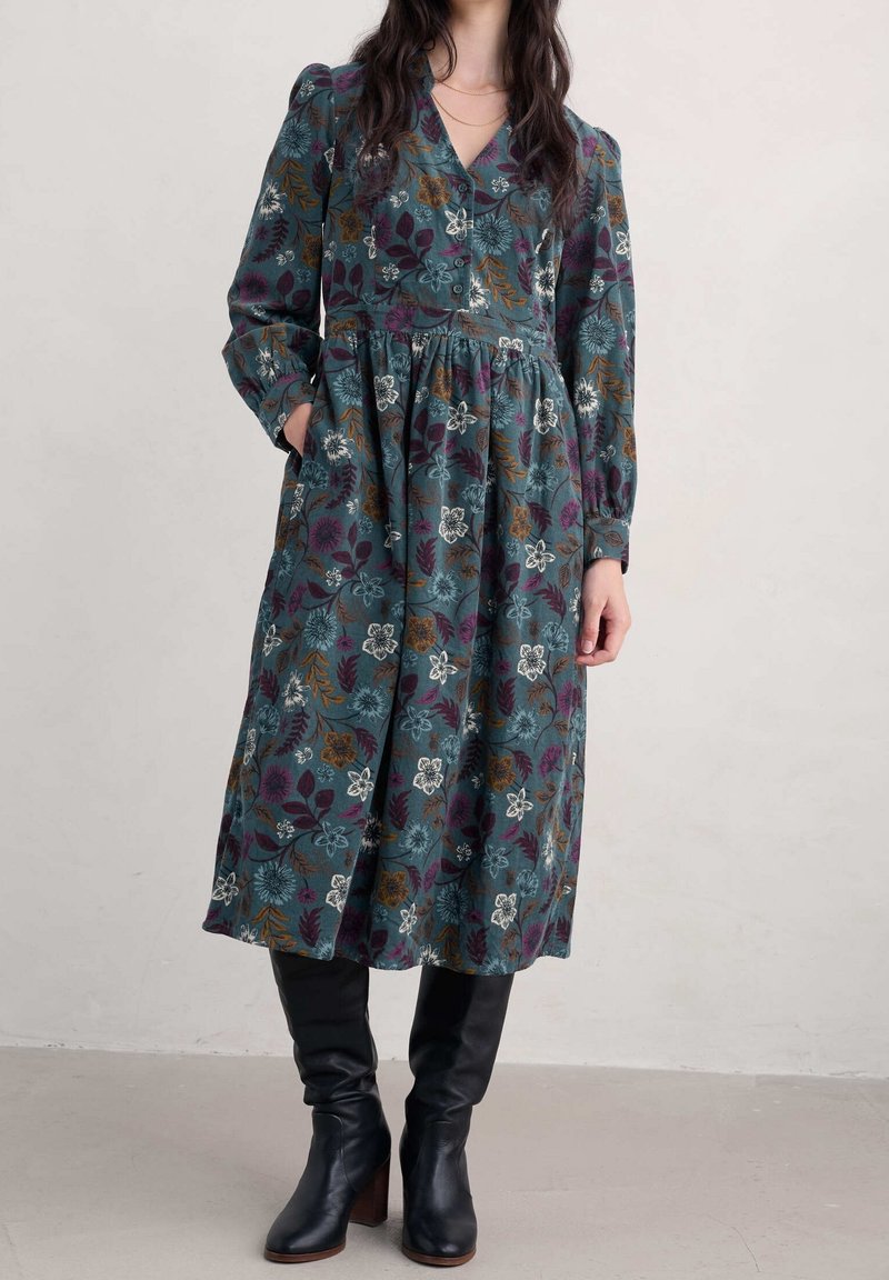 Femme portant une robe fleurie à manches longues et col en V avec des poches et des bottes noires à talons, debout à l’intérieur contre un mur uni.