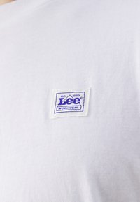 Bílá bavlněná tričko s modrou cedulkou se nápisem "Lee WORKWEAR". Cedulka má obdélníkový tvar s zaoblenými rohy.