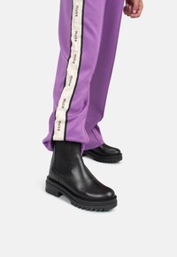 Botas de tobillo de cuero negro con suela gruesa y paneles laterales elásticos, combinadas con pantalones morados que tienen una franja de logo en blanco y negro.
