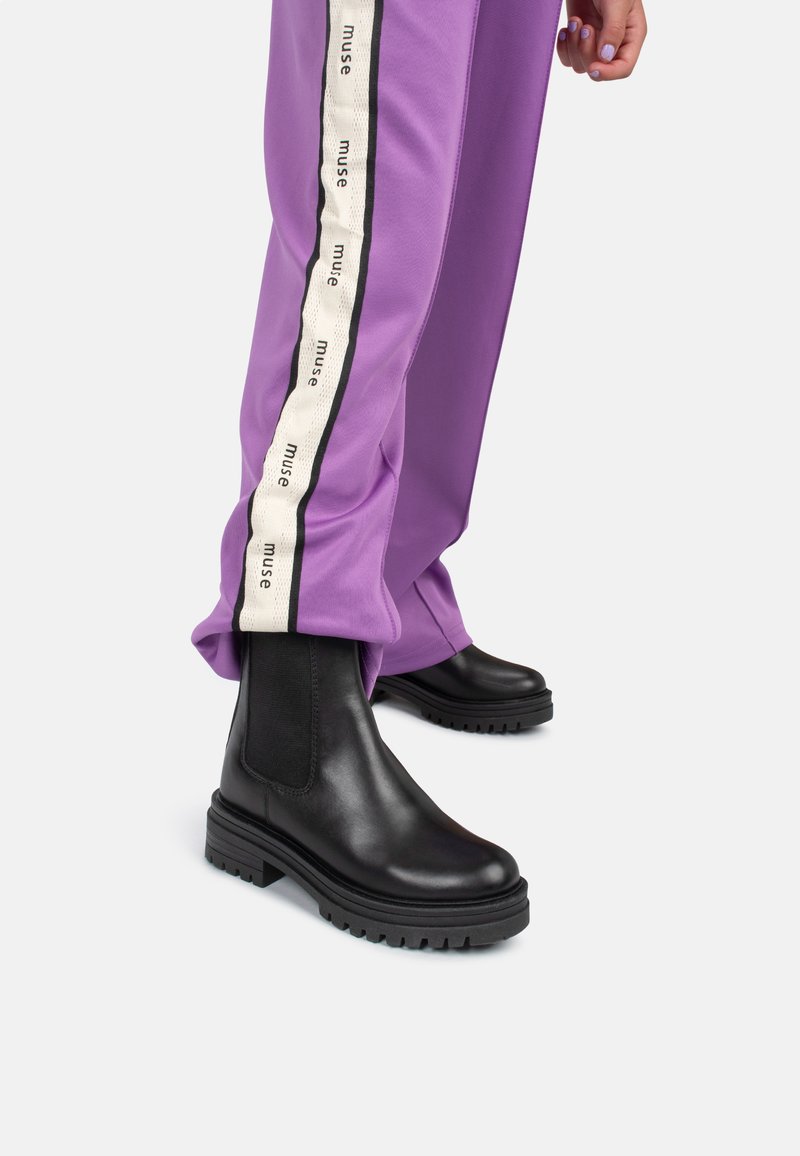 Botas de tobillo de cuero negro con suela gruesa y paneles laterales elásticos, combinadas con pantalones morados que tienen una franja de logo en blanco y negro.