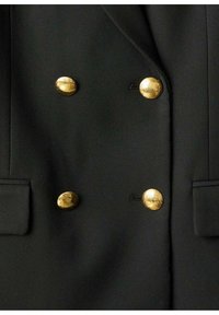Zwarte blazer met dubbelbreasted ontwerp, textuurstof en prominente gouden ronde knopen met een lichte glans. Inclusief pocketdetails.