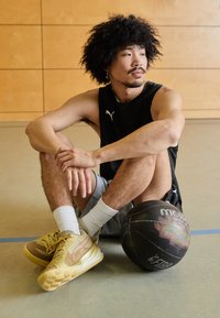 Atleta con top senza maniche nero e pantaloni grigi, seduto con capelli ricci, scarpe da ginnastica gialle e una palla da basket nera accanto a lui.