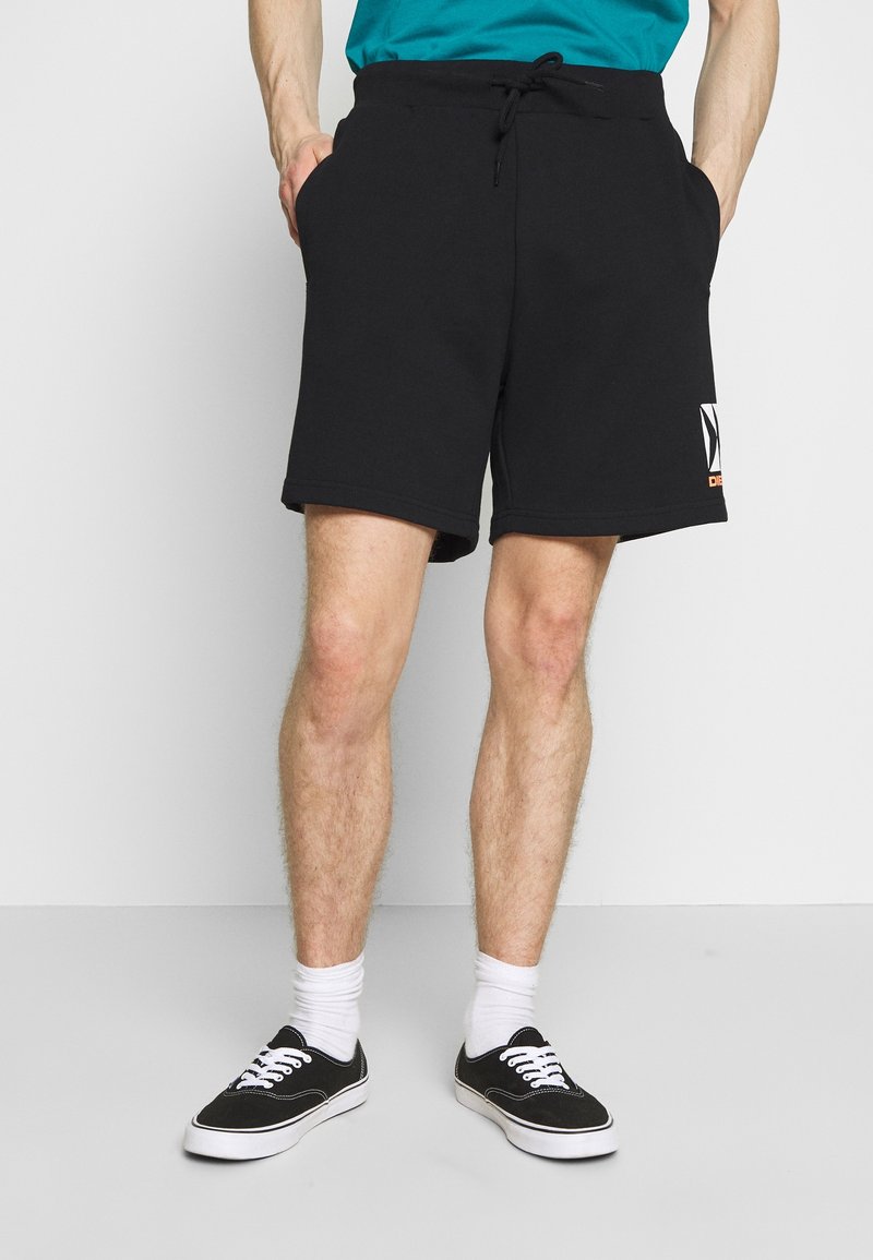 Homme portant un short noir à cordon avec des poches latérales, des chaussettes blanches et des baskets noires basses, debout devant un fond uni.